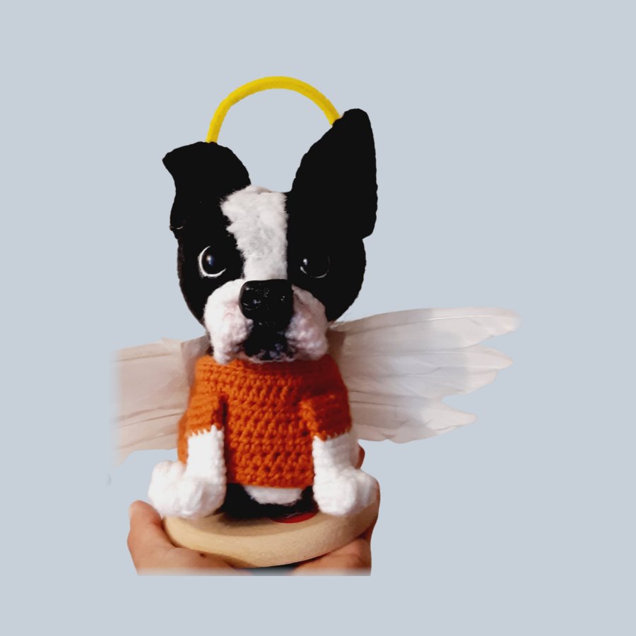 Figura tejida de un perro con suéter naranja, alas y halo sobre una base de madera